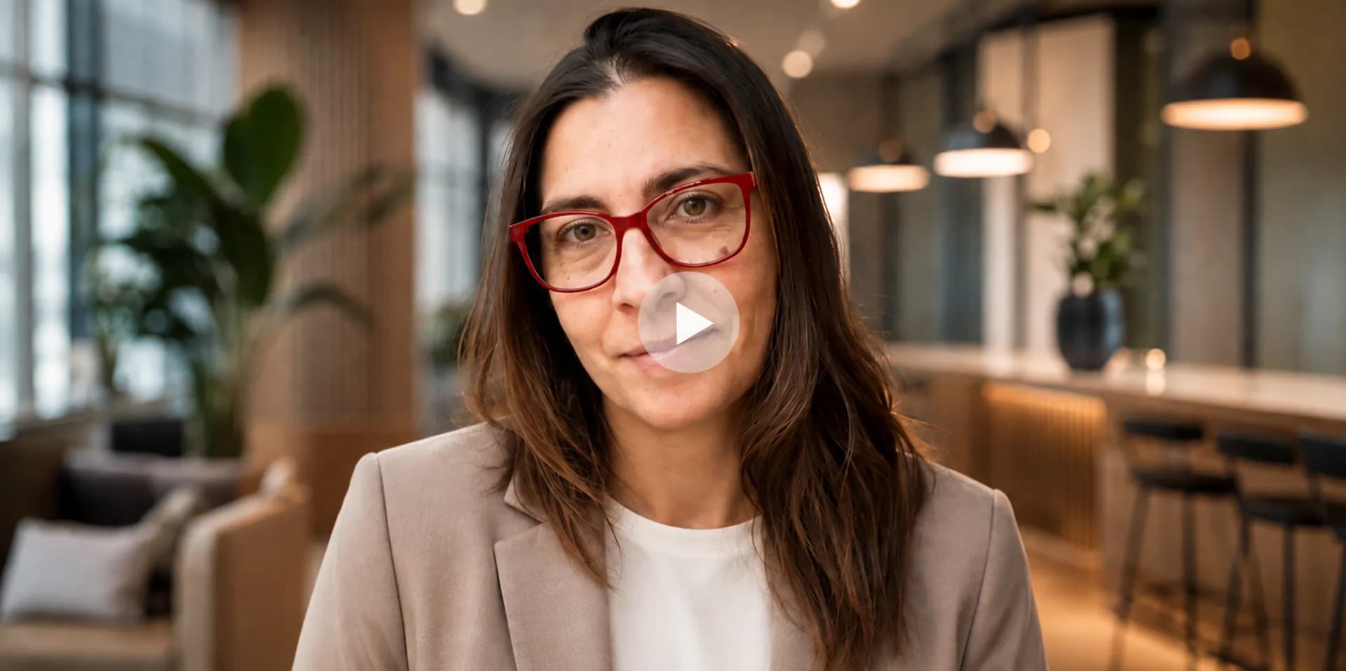 Mujer de gafas rojas y blazer beige mira a cámara en una oficina luminosa; fotograma provisional del video de presentación